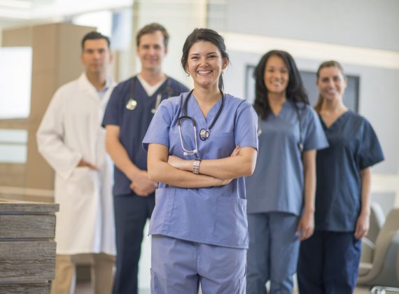 A multi-ethnic group of nurses and a doctor are standing together in their hispital clinic. They are all smiling and looking at the camera.